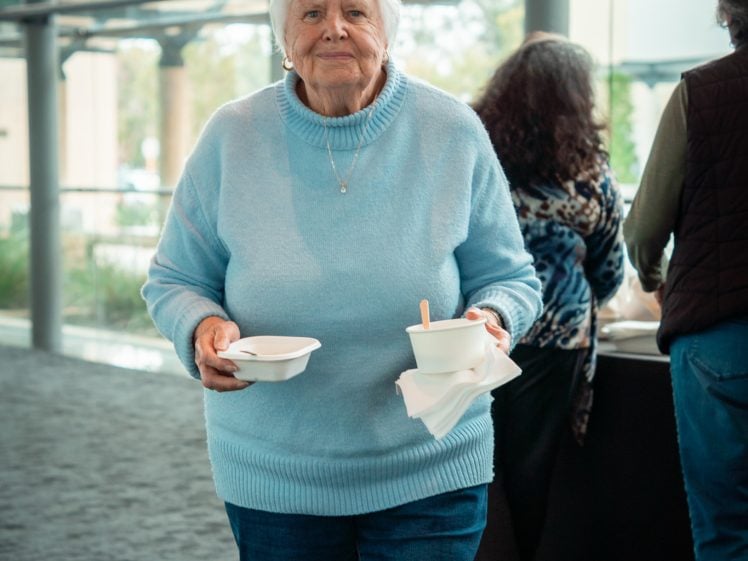 Image of An older woman with short white hair, wearing a light blue jumper and jeans, holds food containers while standing indoors on a carpeted floor. Other people and large windows are visible in the background.