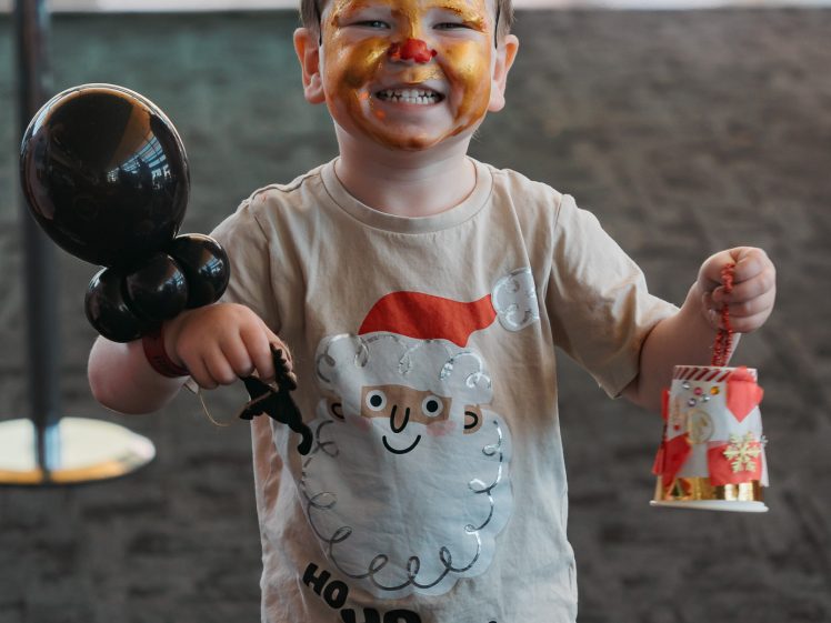 Image of A smiling child with a painted face and blue pipe cleaner antennae wears a Santa t-shirt and red shorts, holding a black balloon figure in one hand and a festive bell-shaped decoration in the other.