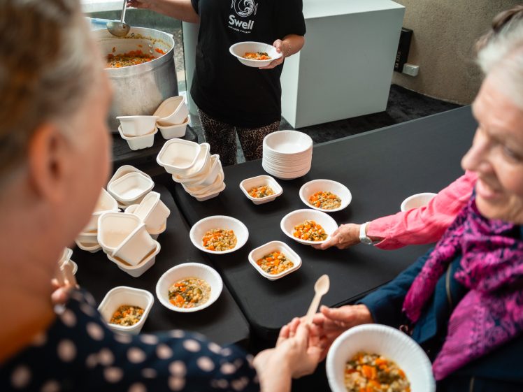 Image of People are being served bowls of vegetable soup from a large pot at a table, with several bowls and containers arranged nearby. Two women in the foreground are receiving their portions and smiling.