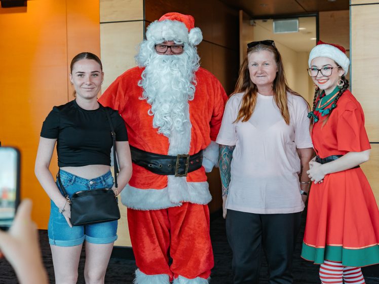 Image of Four people pose together indoors: a woman in a black top and shorts, a person dressed as Father Christmas, a woman in a pink shirt, and a person in a festive red elf costume. A hand holds a mobile to take their photo.