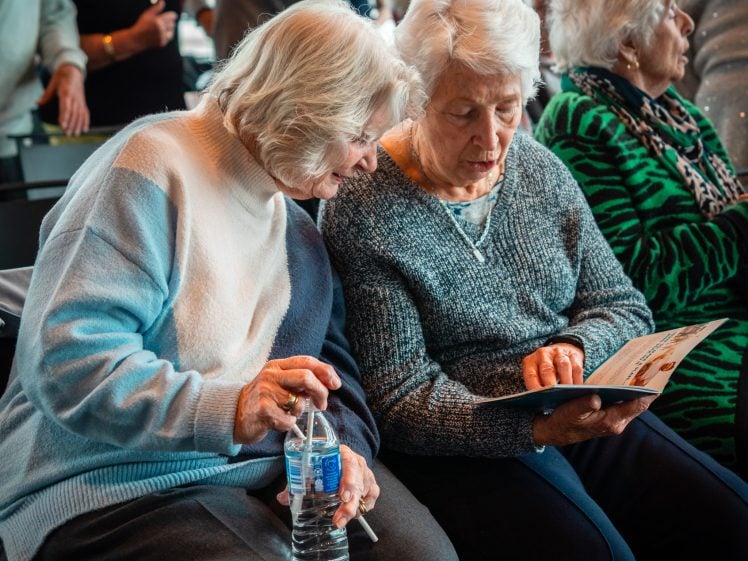 Image of Two elderly women sit closely together; one holds a water bottle while the other points at a leaflet or programme. They appear to be engaged in discussion, surrounded by other pensioners in a brightly lit indoor setting.