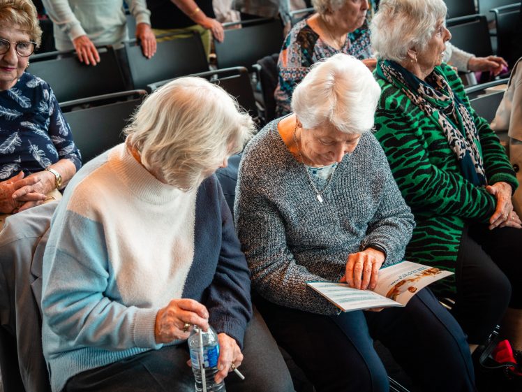 Image of Elderly women sit together in a row of chairs; two in the centre look at a brochure, while others around them chat and wait, creating a friendly, social atmosphere.