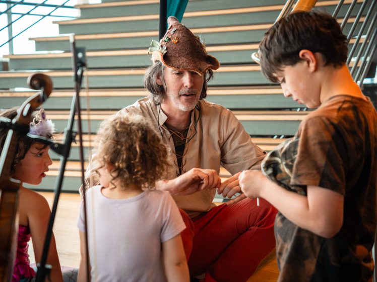 Image of A man in a brown hat and tan shirt kneels and interacts with three children in a bright room with wooden stairs in the background. The children seem engaged and attentive to what he is showing them.