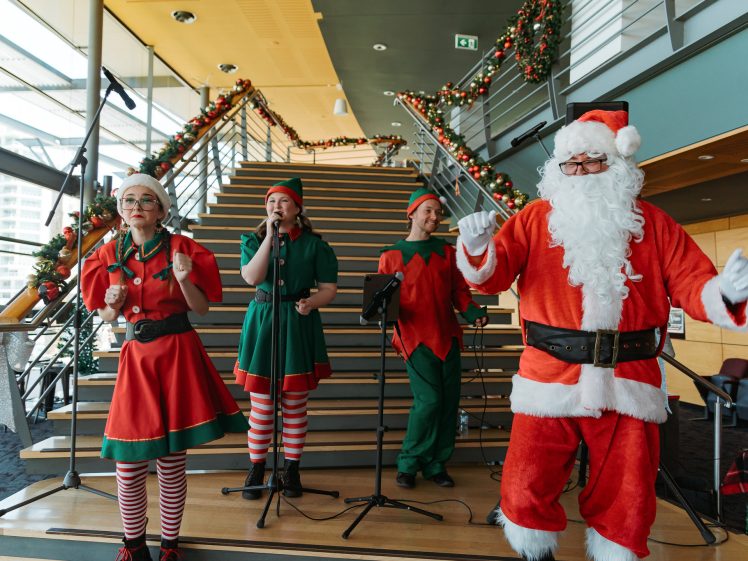 Image of Four people dressed as Father Christmas and elves perform on a decorated staircase indoors, with microphones and festive Christmas garlands on the bannisters.
