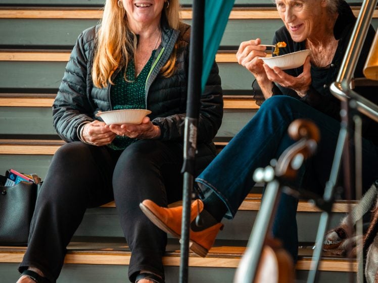 Image of Two older women sit on wooden benches, smiling and eating from bowls. One woman holds a fork mid-mouthful. Musical instruments are partially visible in the foreground. The scene feels casual and friendly.
