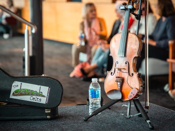 Image of A violin on a stand next to a water bottle and a black instrument case labelled The Jarrah Celts. Four people are seated and talking in the blurred background.