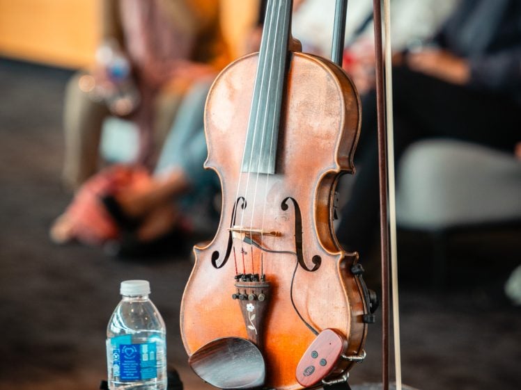 Image of A violin rests on a stand next to a bow and a bottle of water, with a group of blurred people sitting and talking in the background.