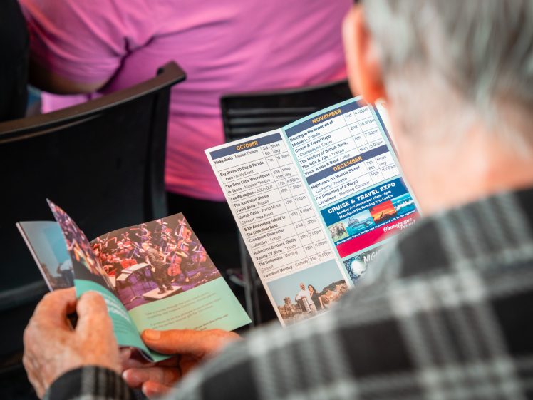 Image of A person in a checked shirt reads an event programme featuring a photo of an orchestra and a schedule of activities, while seated among others at an indoor event.