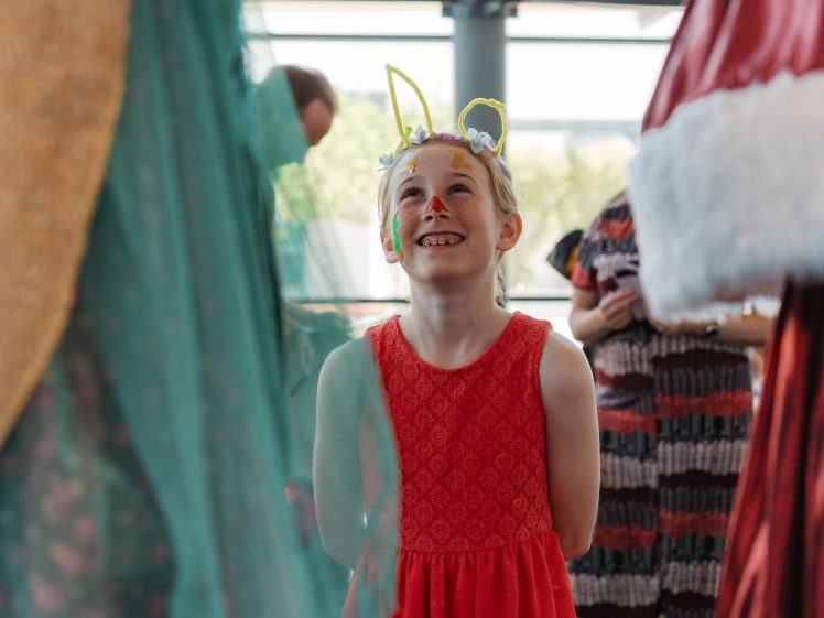 Image of A young girl wearing a red dress, bunny ears, and face paint smiles and looks up, surrounded by people in colourful costumes at what appears to be a festive indoor event.