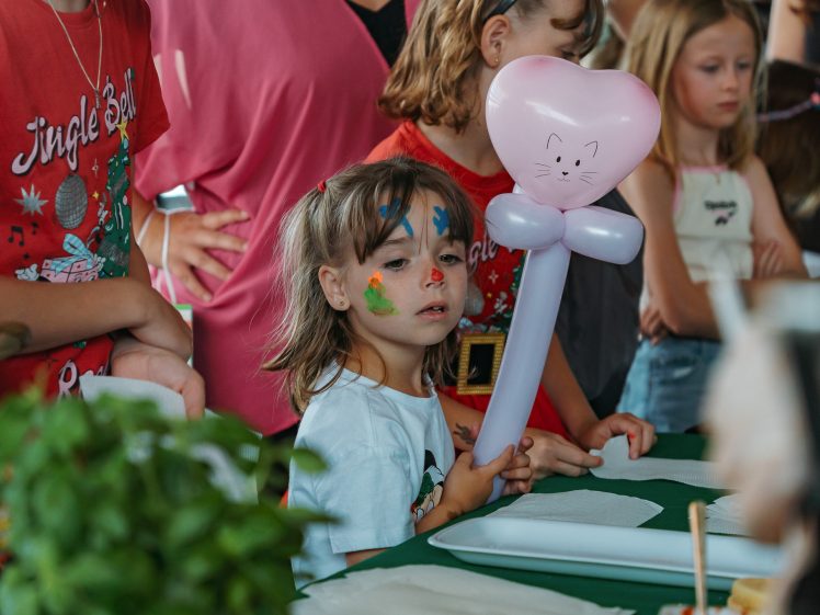 Image of A young child with face paint holds a pink balloon shaped like a cat whilst standing at a table, surrounded by other children and adults at what appears to be a festive event.