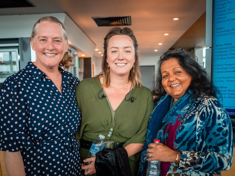 Image of Three women standing together indoors, smiling at the camera. One wears a navy polka dot dress, another a green blouse, and the third has a patterned scarf. They appear to be at a social or professional event.