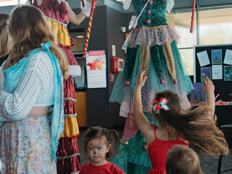 Image of Two performers on stilts dressed in colourful, whimsical costumes entertain a group of children indoors. One performer wears pink pigtails; the other has red curls. The children are smiling and looking up excitedly at the performers.