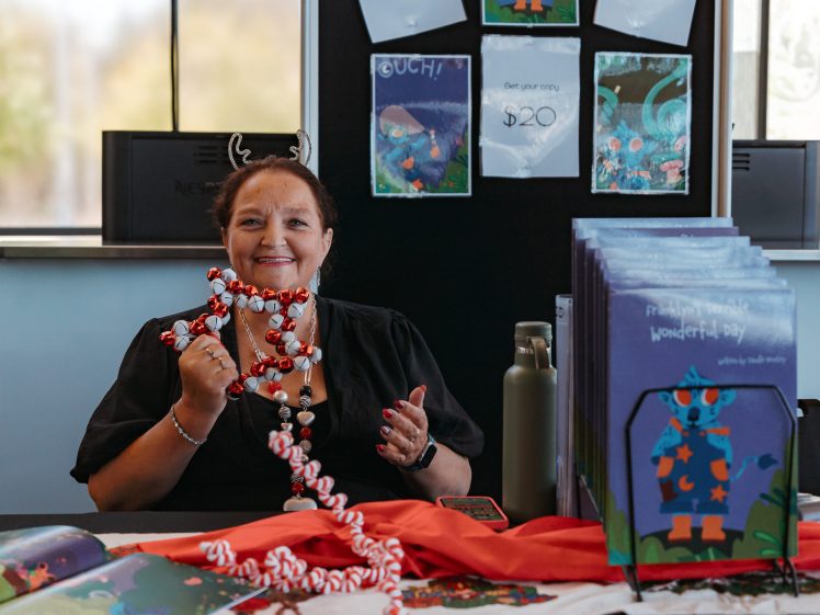 Image of A smiling woman with festive antlers holds a red-and-white beaded garland at a table displaying children's books and decorations. Colourful book covers and signs are visible behind her.