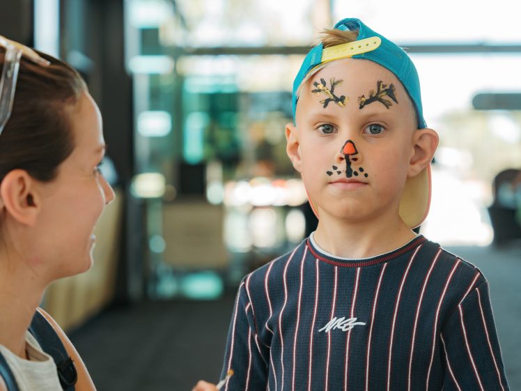 Image of A young boy wearing a blue cap backwards has his face painted like a reindeer. He looks forward with a serious expression whilst an adult woman faces him and smiles in a brightly lit indoor space.