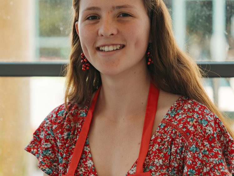 Image of A young woman wearing a red floral dress, red apron, and a festive headband with antler-shaped decorations smiles whilst sitting at a table indoors.
