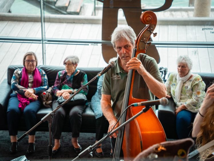 Image of An older man plays a double bass and sings into a microphone indoors, while four older women sit on a sofa in the background, watching and listening. Large windows provide a view of water and a wooden decking outside.