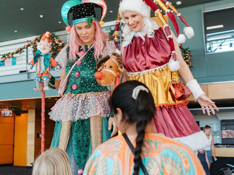 Image of Two performers in colourful, festive costumes and hats stand on stilts, entertaining two children indoors. Holiday decorations are visible in the background.