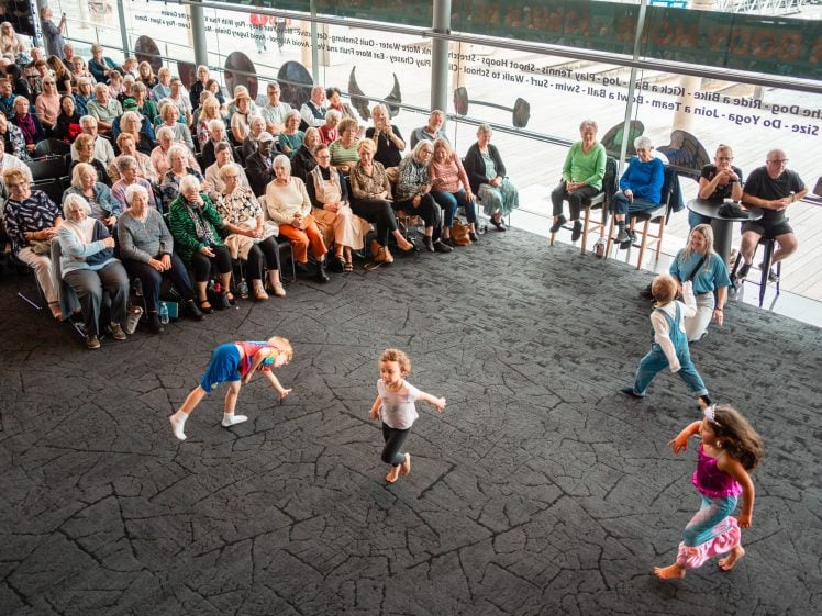 Image of A group of young children dance energetically on a stage in front of a large seated audience of mostly older adults, who watch attentively in a modern, glass-walled venue.