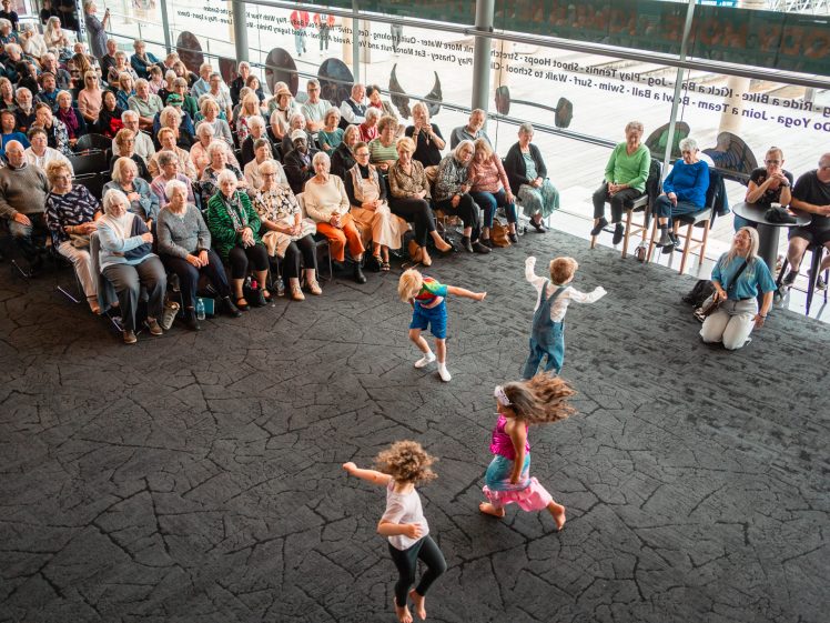 Image of Four young children dance energetically on a black stage in front of a seated audience of older adults in a bright, modern venue with large windows.