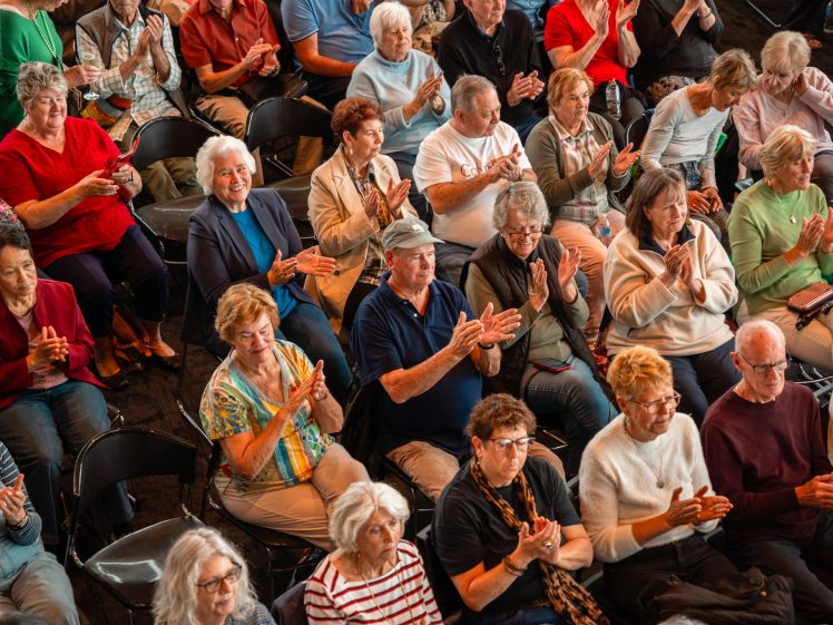 Image of A diverse group of older adults sits in rows of chairs, smiling and enthusiastically applauding, suggesting they are enjoying a live performance or event.