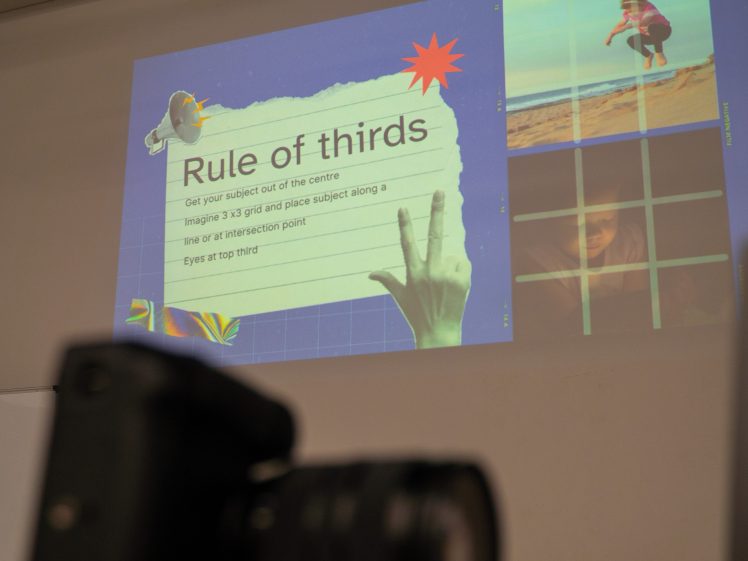 Image of A projected slide titled Rule of thirds explains photography composition tips, with sample images on a grid. A camera is blurred in the foreground.