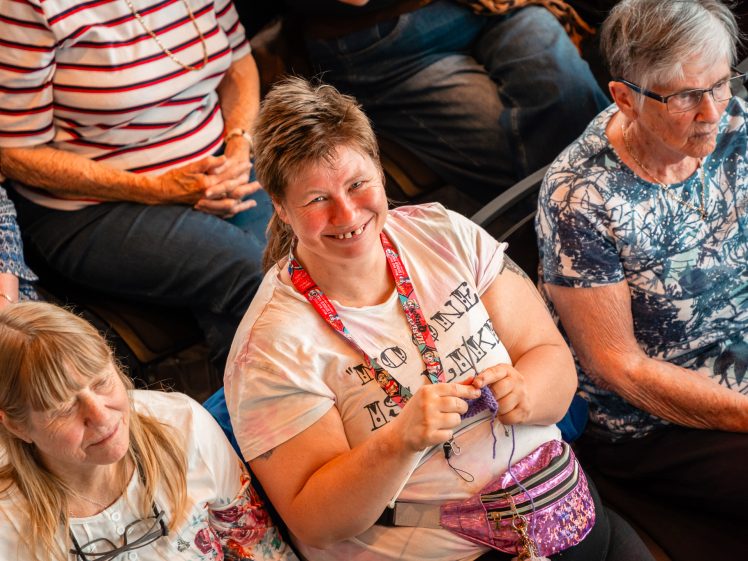 Image of A group of older women sit closely together, some with hands folded. A woman in the centre smiles at the camera, wearing a bum bag and holding wool as if knitting or crocheting. The mood seems cheerful and friendly.