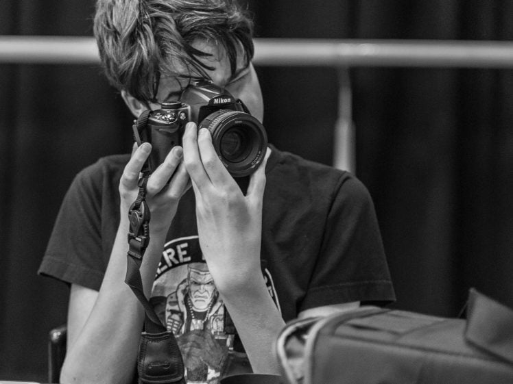 Image of A person with messy hair holds a Nikon camera up to their face, preparing to take a photo. They are seated at a table with a camera bag, lens cap, and papers, against a dark background.