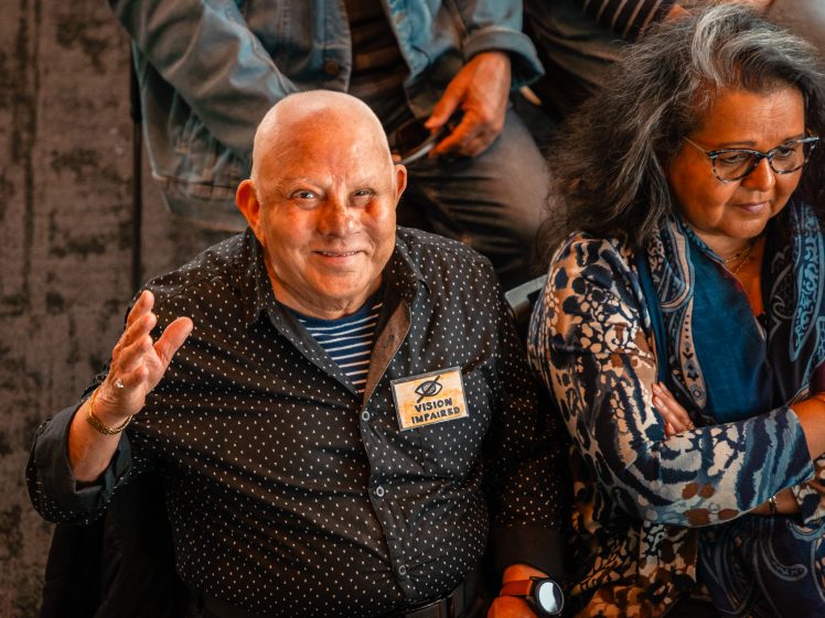 Image of An older man with a bald head and spotty shirt smiles and waves at the camera. He wears a Vision Thailand name badge. A woman with long grey hair and glasses sits beside him with her arms folded, looking down.