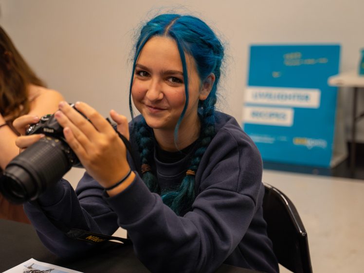 Image of A person with blue plaited hair, wearing a navy sweatshirt, sits at a table holding a Nikon camera and smiling. A blurred blue sign and another person are in the background.