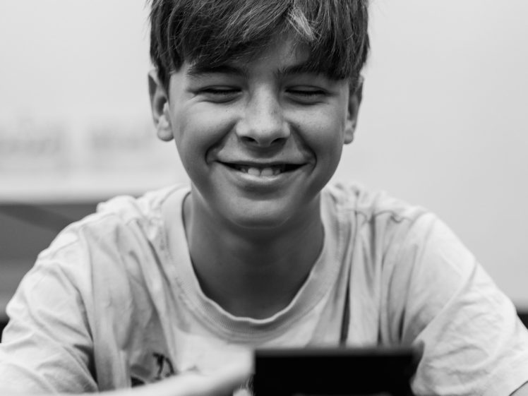 Image of A smiling boy with closed eyes sits at a table, reaching towards a camera in the foreground. The image is in black and white.