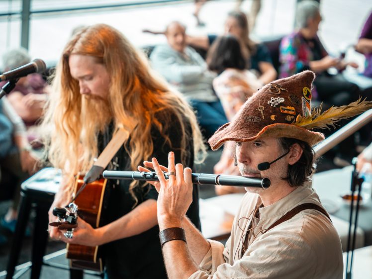 Image of A man wearing a feathered hat plays a flute, whilst another long-haired man plays guitar beside him. Both are performing indoors with a seated audience in the background.