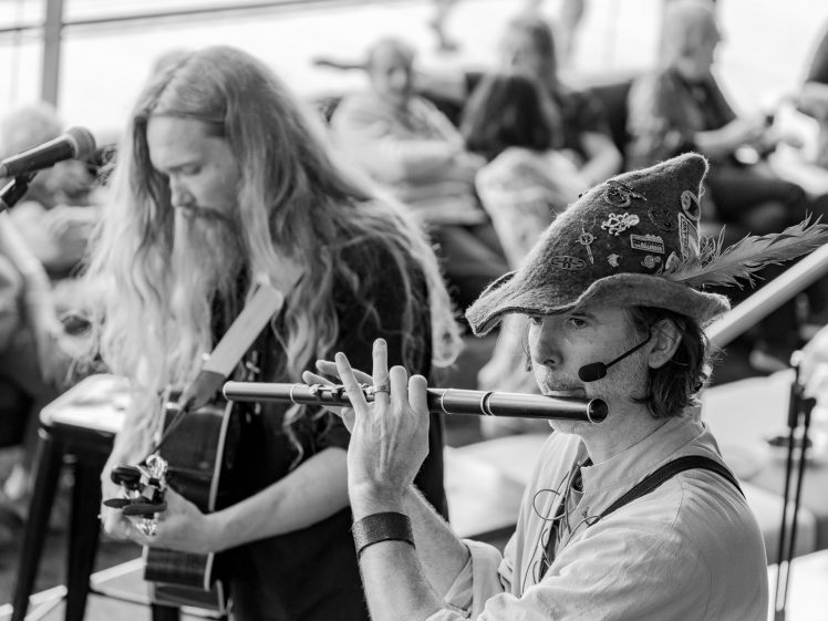 Image of Two musicians perform indoors; one plays a flute and wears a feathered hat with pins, while the other plays guitar. Both have long hair. People are seated and watching in the blurred background. The image is in black and white.