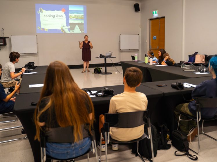 Image of A woman stands in front of a classroom giving a presentation on Leading lines to students seated around three sides of a U-shaped table set-up. Students have notebooks and cameras in front of them.