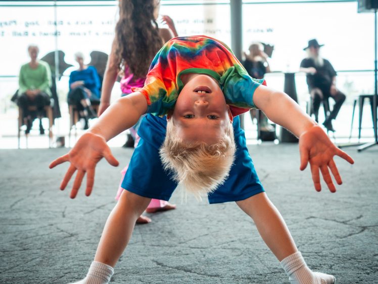 Image of A young child in a colourful tie-dye shirt and blue shorts bends backwards, smiling and looking upside down at the camera, with arms outstretched. Other children and seated adults are visible in the background.