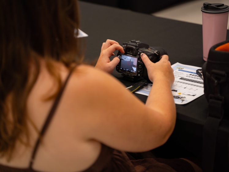 Image of A person sitting at a table adjusts settings on a digital camera. In front of them are a camera instruction manual, a black camera bag, and a pink travel mug. The scene suggests a photography workshop or class.