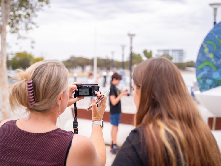 Image of Two women stand outdoors, one holding a camera and taking a photo. They are focused on the camera's screen. The background is slightly blurred, showing a boy, trees, and modern buildings.
