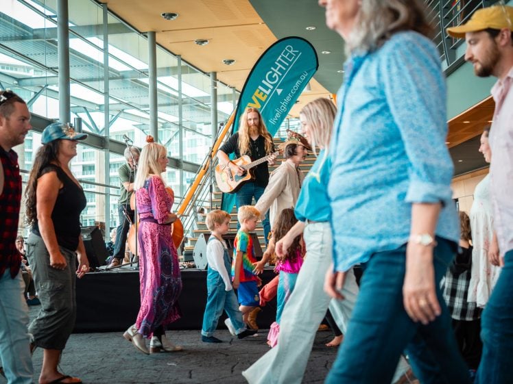 Image of A musician plays guitar on stage as adults and children walk by in a busy indoor space with large windows and modern architecture. A blue Welchester banner is visible behind the performer.