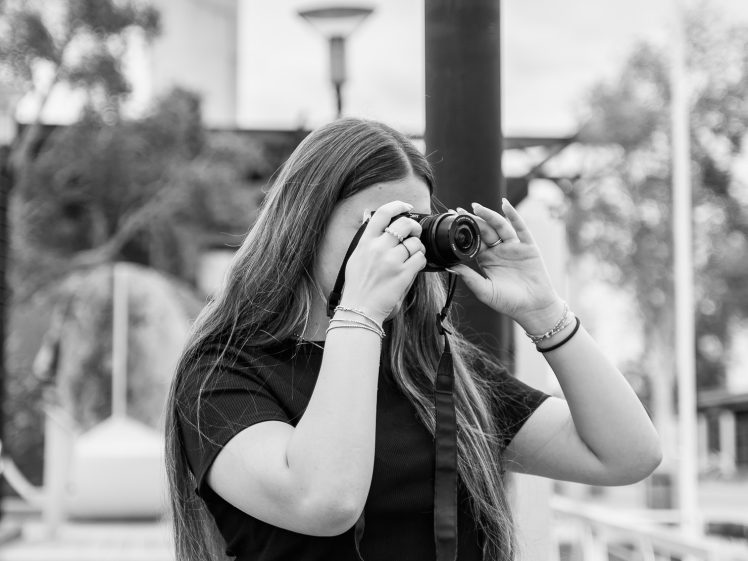 Image of A young woman with long hair, wearing a black crop top and jeans, holds a camera up to her face, taking a photo outdoors on a walkway. The image is in black and white.