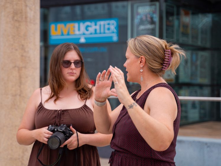 Image of Two women stand outside talking; one holds a camera and listens whilst the other gestures with her hands. Both wear sleeveless dresses. A LiveLighter sign is visible in the background.