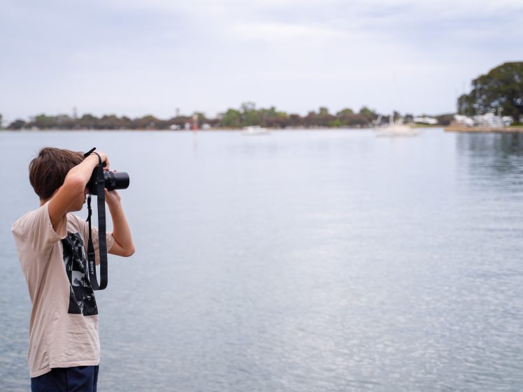 Image of A person with brown hair holds a camera up to their face, taking a photo by a calm lake with trees and boats in the background on a cloudy day.