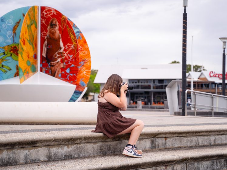 Image of A person in a brown dress sits on outdoor steps, taking a photo with a camera. Behind them is a colourful public artwork with vibrant patterns and a central figure. Buildings and signs are visible in the background.