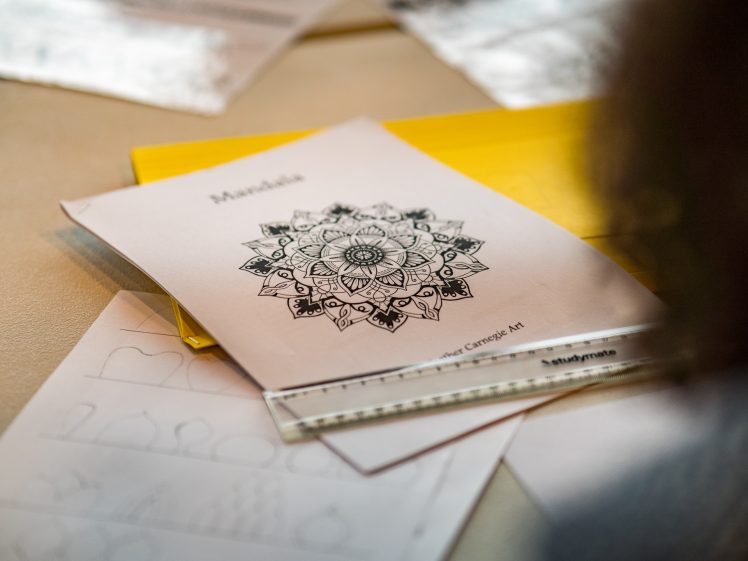 Image of A close-up of a table with a sheet of paper featuring a detailed mandala design, a ruler, and other sheets with geometric sketches, suggesting an art or drawing activity in progress.