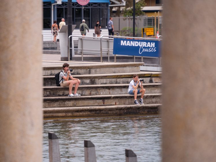 Image of Two boys sit on steps by the water, separated by a distance, both looking at their mobiles. In the background, people walk near a building with a Mandurah Cruises sign. The image is framed by two pillars.