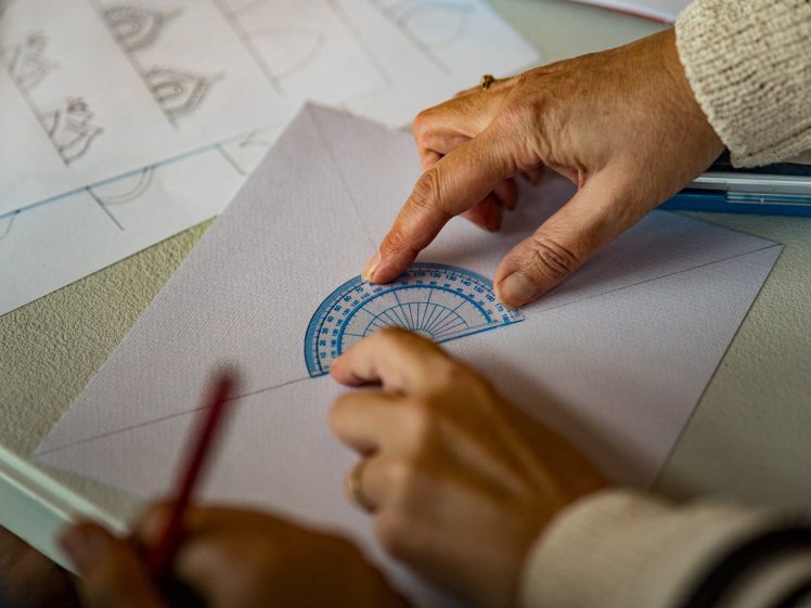 Image of Two people use a blue protractor to measure or draw an angle on a white sheet of paper. One person holds the protractor while the other draws with a red pencil. Sketches and papers are visible in the background.