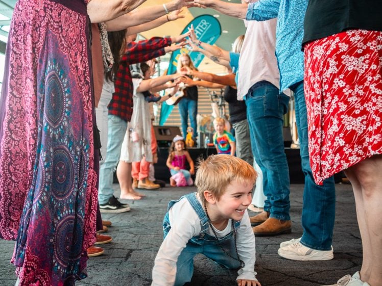 Image of A young child crawls under an archway formed by the raised arms of smiling adults standing in two queues, engaging in a playful group activity indoors.
