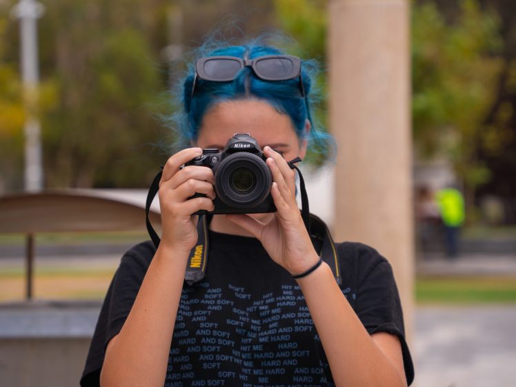 Image of Person with blue hair and sunglasses on their head holds a Nikon camera up to their face, taking a photo outdoors. They wear a black T-shirt, and the background is blurred with trees and columns visible.