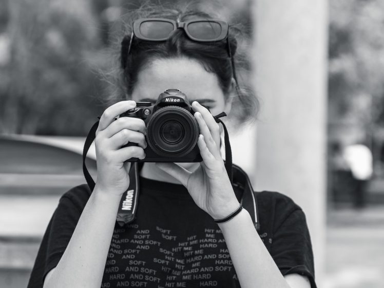 Image of A person with sunglasses on their head holds a Nikon camera, taking a photo. The image is black and white, and the person is wearing a dark shirt with text. The background is outdoors and blurred.