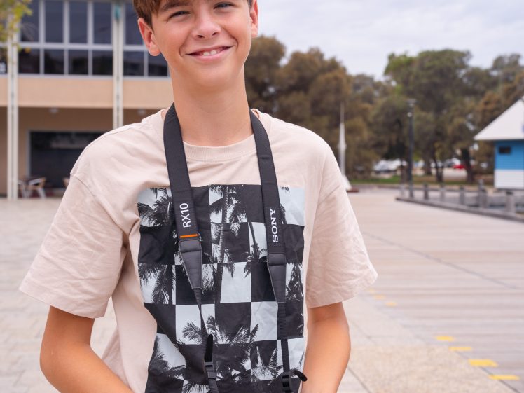Image of A teenage boy with brown hair, wearing a beige t-shirt with a black and white graphic, stands outdoors holding a Sony camera and smiling at the camera. Buildings and trees are visible in the background.