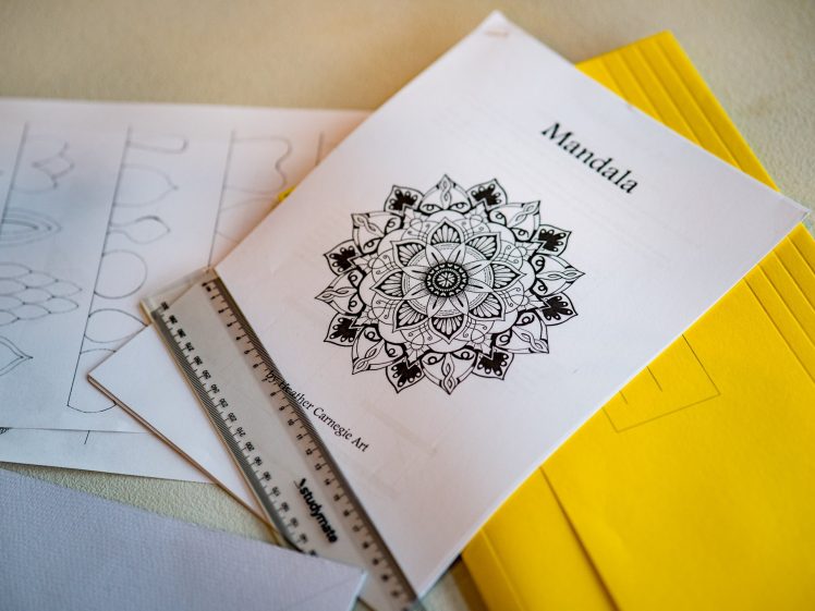 Image of A printed mandala design on white paper sits under a ruler, next to yellow envelopes and sketch sheets with geometric patterns, on a table.