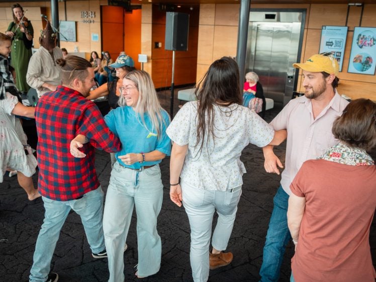 Image of A group of adults dance together in a circle inside a modern building. They are holding hands and smiling, whilst others watch and join in the background. The atmosphere appears lively and joyful.